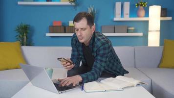 Man using laptop at home looking at his phone, texting, not wanting to work. Man working from home on laptop, looking at him with phone in hand, surfing social media, texting. video