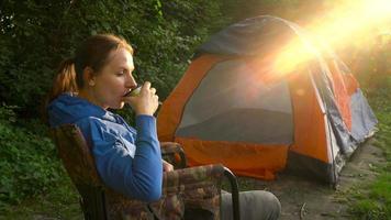 vrouw is drinken thee in een fauteuil in de buurt een tent Aan de rivier- bank video