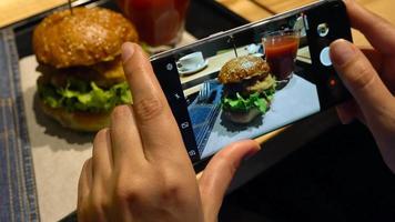 Girl makes a photo of burger and tomato juice on a smartphone in a cafe close up video