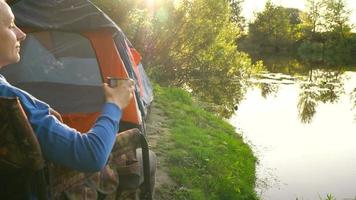 vrouw is drinken thee in een fauteuil in de buurt een tent Aan de rivier- bank video