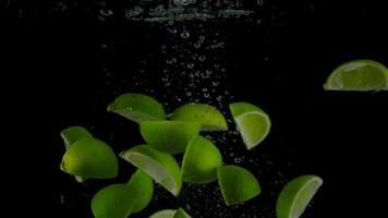 Lime pieces fall and float in water, black background. Slow motion video