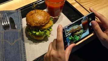 meisje maakt een foto van hamburger en tomaat sap Aan een smartphone in een cafe dichtbij omhoog video