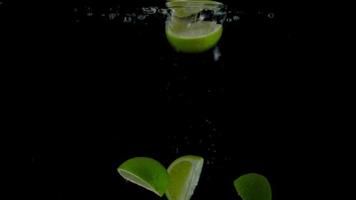 Lime pieces fall and float in water, black background. Slow motion video