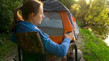 vrouw is drinken thee in een fauteuil in de buurt een tent Aan de rivier- bank video