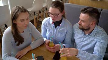 Three friends sit in a cafe, drink juice and have fun communicating video