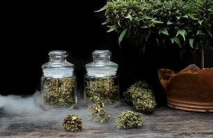 The marijuana buds in a clear glass jar sit on a smoky floor at the black background photo