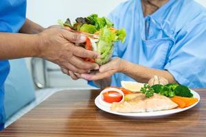 asiático mayor mujer paciente comiendo salmón filete desayuno con vegetal sano comida mientras sentado y hambriento en cama en hospital. foto