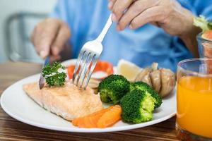 Asian senior woman patient eating Salmon steak breakfast with vegetable healthy food while sitting and hungry on bed in hospital. photo