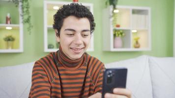 Happy boy at home using phone, texting, girlfriend, in good spirits. Boy spending time with his smartphone at home, texting, social media, mobile games, dating sites. video