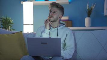 jong Mens bedachtzaam komt eraan omhoog met creatief en ingenieus idee. jong Mens zittend Aan sofa Bij huis gebruik makend van laptop denken en vinden een mooi zo idee en wezen gelukkig. video