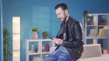 elegante e freddo vestito adulto giovane uomo sms su il Telefono felicemente e emotivamente. contento e bello giovane uomo sms, sorridente, godendo utilizzando il suo smartphone a casa. video