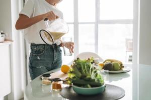 Young slim woman in white t-shirt and blue jeans pouring fruit smoothie healthy food in kitchen at home photo