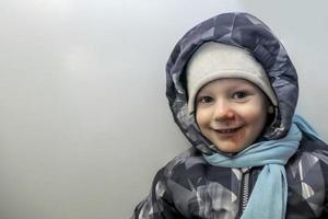 contento chico en un gris mono, un sombrero y un azul bufanda con abrasiones ungido con yodo en su rostro. niño lesión. bandera con un Copiar espacio foto