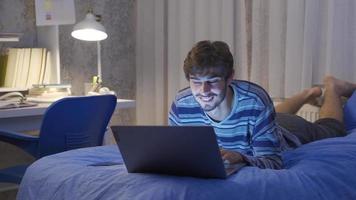 Young man lying in bed looking at laptop. Young man working on computer lying in bed at night. Happy and motivated. video