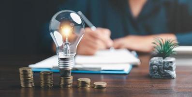 Businesswoman with lightbulb on coins and using calculator to calculate and money stack. Save energy and money with accounting finance in office concept. Idea of energy saving planning in home. photo