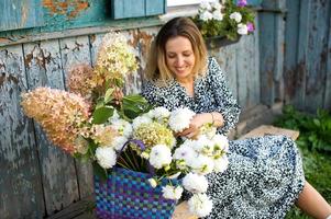 A cute girl is sitting on a bench with a basket of dahlias and hydrangeas in the village photo