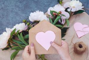 Female hands hold envelope with pink paper heart, sending flowers and invitation for romantic event photo