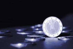 One euro coin standing on edge shining in spotlight against many different coins on dark background. Money flow photo