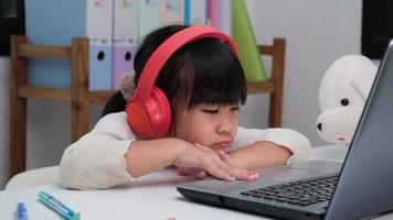 Cute elementary school girl wearing headphones and using a laptop computer. Happy Asian kids study online interactively with laptop computer or homeschooling, listening to music or playing games. video