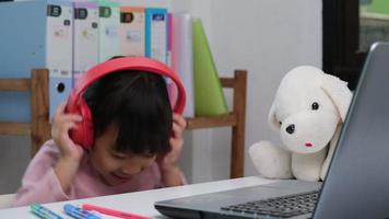 carino elementare scuola ragazza indossare cuffie e utilizzando un' il computer portatile computer. contento asiatico bambini studia in linea interattivamente con il computer portatile computer o istruzione a casa, ascoltando per musica o giocando Giochi. video