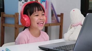fofa elementar escola menina vestindo fones de ouvido e usando uma computador portátil computador. feliz ásia crianças estude conectados interativamente com computador portátil computador ou Educação escolar em casa, ouvindo para música ou jogando jogos. video
