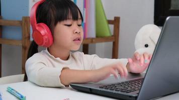 fofa elementar escola menina vestindo fones de ouvido e usando uma computador portátil computador. feliz ásia crianças estude conectados interativamente com computador portátil computador ou Educação escolar em casa, ouvindo para música ou jogando jogos. video