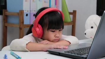 Cute elementary school girl wearing headphones and using a laptop computer. Happy Asian kids study online interactively with laptop computer or homeschooling, listening to music or playing games. video