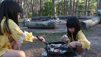 Happy family, cute sisters sitting on picnic by stove near tent and barbecuing in pine forest. Happy family on vacation in nature. video