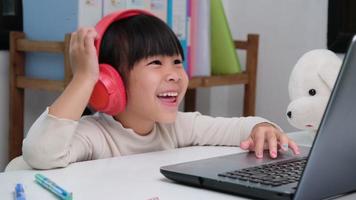 Cute elementary school girl wearing headphones and using a laptop computer. Happy Asian kids study online interactively with laptop computer or homeschooling, listening to music or playing games. video