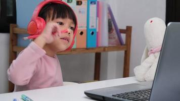 linda elemental colegio niña vistiendo auriculares y utilizando un ordenador portátil computadora. contento asiático niños estudiar en línea interactivamente con ordenador portátil computadora o educación en el hogar, escuchando a música o jugando juegos. video