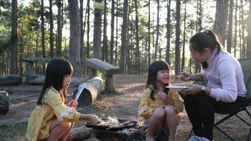 Happy mother and children picnic sit by stove near tent and grill a barbecue in pine forest, eating and have conversation. Happy family on vacation in nature video