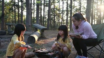 Happy mother and children picnic sit by stove near tent and grill a barbecue in pine forest, eating and have conversation. Happy family on vacation in nature video
