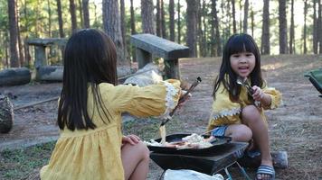 Happy family, cute sisters sitting on picnic by stove near tent and barbecuing in pine forest. Happy family on vacation in nature. video