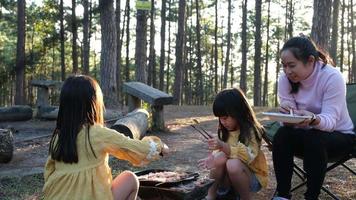 Happy mother and children picnic sit by stove near tent and grill a barbecue in pine forest, eating and have conversation. Happy family on vacation in nature. video