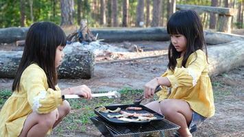 Happy family, cute sisters sitting on picnic by stove near tent and barbecuing in pine forest. Happy family on vacation in nature. video