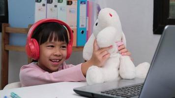 Cute elementary school girl wearing headphones and using a laptop computer. Happy Asian kids study online interactively with laptop computer or homeschooling, listening to music or playing games. video