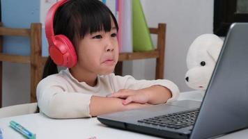 Cute elementary school girl wearing headphones and using a laptop computer. Happy Asian kids study online interactively with laptop computer or homeschooling, listening to music or playing games. video