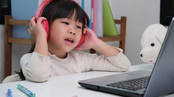 mignonne élémentaire école fille portant écouteurs et en utilisant une portable ordinateur. content asiatique des gamins étude en ligne interactivement avec portable ordinateur ou l'école à la maison, écoute à la musique ou en jouant Jeux. video