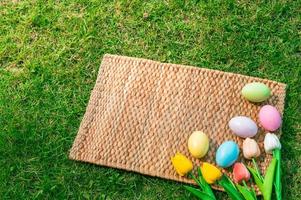 Happy Easter holiday greeting card concept. Colorful Easter Eggs and spring flowers on Papyrus rattan weave texture background. Flat lay, top view, copy space. photo