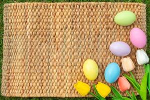 Happy Easter holiday greeting card concept. Colorful Easter Eggs and spring flowers on Papyrus rattan weave texture background. Flat lay, top view, copy space. photo