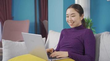 schattig Aziatisch vrouw is aan het kijken film Aan laptop terwijl genieten van en lachend. mooi Aziatisch vrouw zittend alleen in leven kamer aan het kijken film gebruik makend van laptop gelukkig en lachend. video