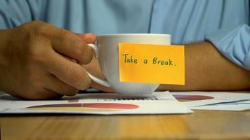 Hand man holding a coffee cup and put it down on the Annual report on coffee cup have a sticky note that says take a break. The concept of encouragement for colleagues video