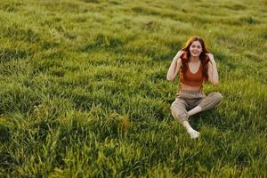 Young woman with red hair is happy and smiling sitting on the fresh grass in the park in the sun photo