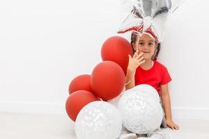 Happy brightful image of cute joyful little girl in tulle skirt sitting on present with balloons isolated on white background. Amazing charming birthday fashionable kid looking to camera photo