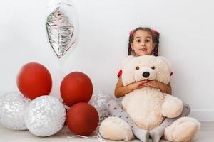 Cute kid little girl posing with red heart shaped balloons and a teddy bear isolated on white. Children fashion photo