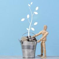 A wooden dummy and a miniature bucket with metal coins and a painted tree with leaves on a blue background. Income and profit growth concept photo