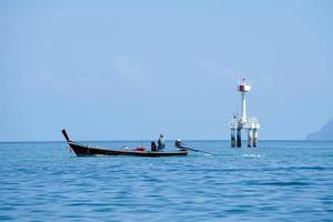 Rear view of fisherman with the passenger drives the long tail boat in the sea with defocused light house in background. The skyline of seascape with blue sky background photo