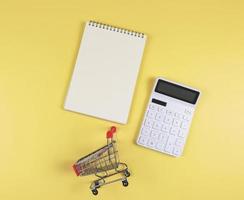 flat layout of opened blank page notebook with shopping trolley or shopping cart and white calculator on yellow background. financial plan concept. photo