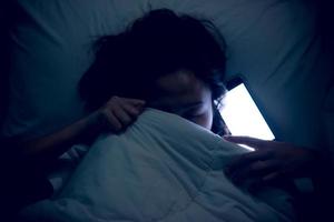 asiático niño jugando juego en teléfono inteligente en el cama a noche, la niña adicto social medios de comunicación foto