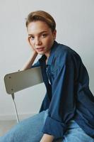 A young woman sitting in a chair at home smiling with teeth with a short haircut in jeans and a denim shirt on a white background. Girl natural poses with no filters photo